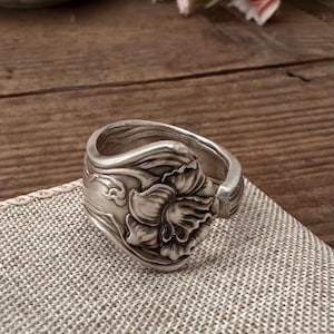 May include: A silver-toned ring with a floral design. The ring features a detailed, raised flower motif on the face, with intricate patterns along the band. The ring is set on a textured, light-colored surface.