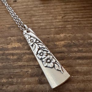 May include: A silver-toned pendant necklace featuring a tapered, flat metal piece with an embossed floral pattern of three flowers and leaves. The pendant is attached to a delicate, thin silver-toned chain link necklace.