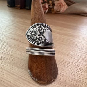 May include: A silver-toned spoon ring featuring an embossed floral pattern with three distinct daisy-like flowers on the wider end and a smooth, multi-grooved band that wraps around the finger. The metal has a polished finish with darkened accents.