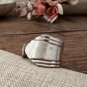 May include: A silver ring made from repurposed silverware. The ring has a layered design with a wide band and a textured surface. The ring is displayed on a neutral-colored fabric.