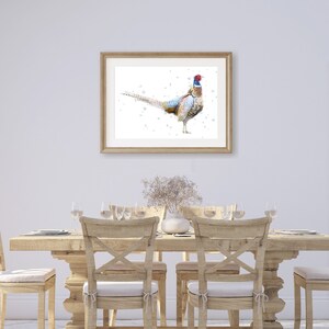 May include: A framed watercolor painting of a pheasant with a white background. The pheasant is standing on a white surface with its tail feathers spread out. The painting is hanging on a white wall in a dining room setting with a wooden table and chairs.