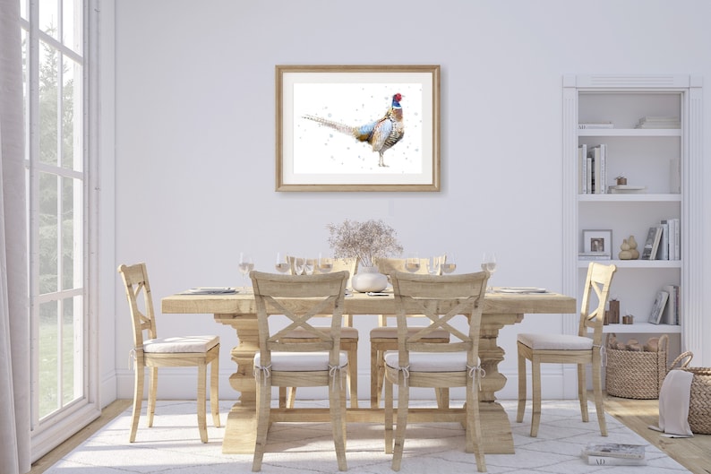 May include: A framed watercolor painting of a pheasant with a white background. The pheasant is standing on a white surface with its tail feathers spread out. The painting is hanging on a white wall in a dining room setting with a wooden table and chairs.