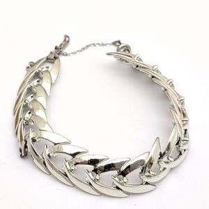 May include: Silver-toned bracelet with a leaf-like design. The bracelet features a series of interconnected, curved metal pieces, creating a textured and eye-catching accessory. A small safety chain is attached for added security. The bracelet is set against a white background.