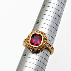 May include: A gold ring with a large, deep red gemstone surrounded by smaller, clear gemstones. The ring is on a metal ring sizer, with the size marked as 9.