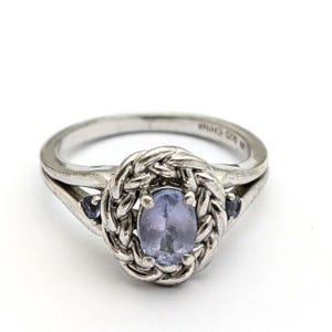 May include: A silver ring with a braided band and a light blue oval-shaped gemstone in the center. Two smaller light blue gemstones are set on either side of the center stone.