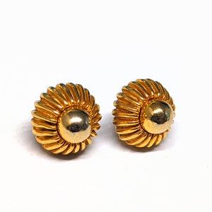 May include: A pair of gold-toned earrings with a textured, circular design. The earrings have a raised center and a slightly domed shape.