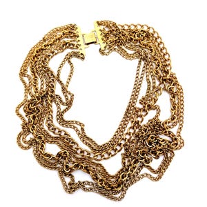 May include: A gold-toned necklace featuring multiple strands of varying chain styles. The necklace has a clasp closure and is displayed against a white background. The chains are layered, creating a bold and textured look. The jewelry is likely vintage or costume.