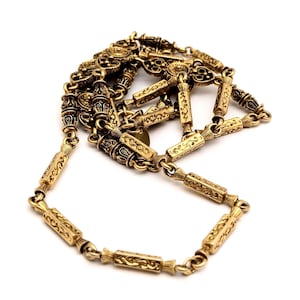 May include: A gold-toned metal necklace featuring a series of rectangular, engraved bar links connected by circular jump rings. The bar links have a textured, ornate pattern, and the chain is arranged in a loose, coiled formation.