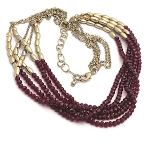 May include: A multi-strand necklace featuring alternating sections of gold-toned, elongated beads and strands of small, faceted, deep red beads. The necklace has a chain closure and an adjustable length.
