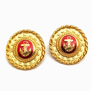 May include: Pair of gold-toned, round earrings with a nautical theme. Each earring features a red center with a gold anchor design, surrounded by a gold rope-like border. The outer ring has embossed letters. The earrings are set against a white background.