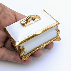 May include: A small white porcelain trinket box with gold accents. The box has a decorative gold handle and sits on four gold feet.