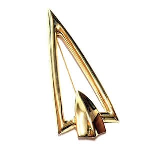 May include: A gold-toned brooch in a geometric design. The brooch features a triangular frame with a pointed end and a smaller, solid, arrow-shaped element. A pin is attached to the back for securing the brooch.
