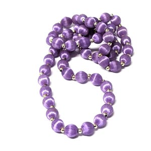 May include: A necklace composed of numerous lavender-colored, spherical beads. Each bead is wrapped in a textured fabric and separated by small, silver-colored metal accents. The necklace is displayed against a white background.