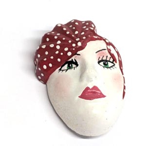 May include: A ceramic face sculpture with a red polka-dotted head covering and red lips. The face has green eyes, rosy cheeks, and is painted on a white background. The sculpture is a decorative art piece.