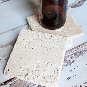 May include: Square, light beige stone coasters with a textured surface. A brown glass bottle rests on one of the coasters. The coasters are made of natural stone and are designed to protect surfaces from water rings.
