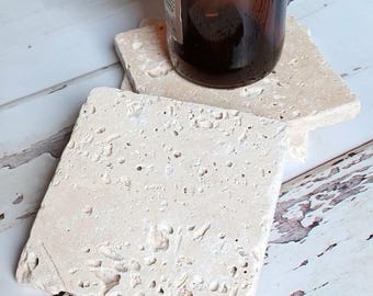 Op de afbeelding: Vierkante, lichtbeige stenen onderzetters met een textuur. Een bruine glazen fles staat op een van de onderzetters. De onderzetters zijn gemaakt van natuursteen en beschermen oppervlakken tegen waterkringen.