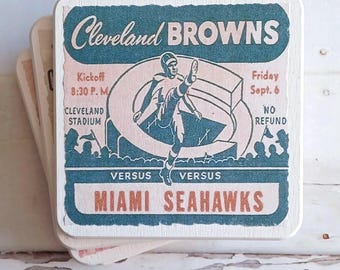 May include: A square coaster featuring a vintage-style graphic of a Cleveland Browns football game. The design includes a player kicking a ball, the text Cleveland BROWNS, Kickoff 8:30 P.M., Friday Sept. 6, CLEVELAND STADIUM, NO REFUND, and MIAMI SEAHAWKS.