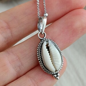May include: A silver chain necklace with a white cowrie shell pendant. The shell is surrounded by a silver border and has a silver bead at the bottom.