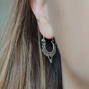 Puede incluir: Pendientes de aro de plata con un diseño delicado y ornamentado. Los pendientes tienen un pequeño encanto colgante en la parte inferior.