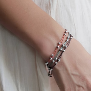 以下が含まれることがあります： 銀色のビーズとカラフルな紐でできた重ね付けブレスレットのセット。ブレスレットは手首につけられています。