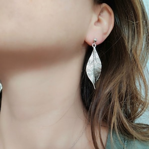 Puede incluir: Pendientes de hoja plateados con forma de hoja y superficie texturizada. Los pendientes los lleva una persona con el pelo largo y castaño.