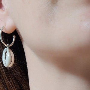 Può includere: Primo piano del collo e delle spalle di una persona che indossa un paio di orecchini a conchiglia d'argento. Gli orecchini sono semplici ed eleganti, con un piccolo ciondolo a conchiglia appeso a ogni cerchio.