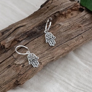 Puede incluir: Pendientes de aro de plata con un dije de mano de hamsa. La mano de hamsa es un símbolo de protección y buena suerte.