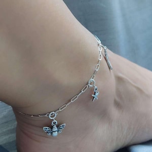 May include: Silver-toned anklet featuring a bee charm, a starfish charm, and a small bar charm. The chain is a link style. The anklet is worn on an ankle.
