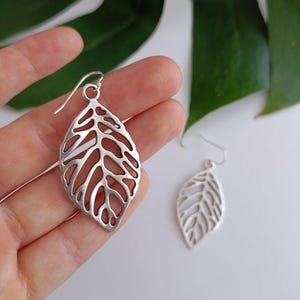 Könnte beinhalten: Ein Paar silberne, blattartige Ohrhänger. Die Ohrringe sind aus Metall und haben ein zartes, filigranes Design.