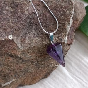 Op de afbeelding: Een zilveren ketting met een paarse amethist punt hanger. De hanger is driehoekig en heeft een gladde, gepolijste afwerking.