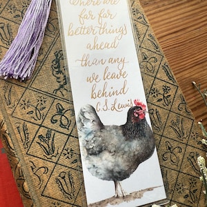 May include: A bookmark with a watercolor illustration of a chicken and the quote "There are far far better things ahead than any we leave behind" by C.S. Lewis. The bookmark has a purple tassel and is on top of a book.