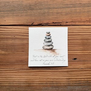 May include: A watercolor painting of a stack of gray stones with a white background. The text "Trust in the Lord with all your heart and lean not on your own understanding. Proverbs 3:5" is written below the stones.