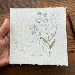May include: A square piece of textured white paper featuring a watercolor illustration of light blue forget-me-not flowers with green stems and leaves. The text reads, "Love each other as I have loved you." John 15:12, written in black ink.