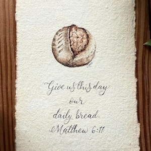 May include: Watercolor painting of a loaf of bread, with a detailed depiction of the crust and interior. The artwork includes the text "Give us this day our daily bread. Matthew 6:11" in elegant script on textured paper.