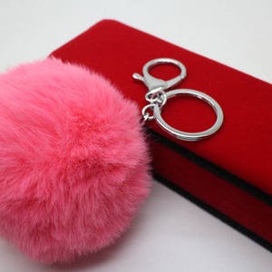 May include: A pink fluffy pom-pom keychain attached to a silver key ring. The keychain is sitting on a red velvet box.