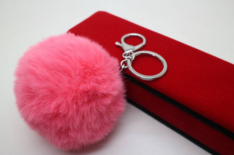 May include: A pink fluffy pom-pom keychain attached to a silver key ring. The keychain is sitting on a red velvet box.