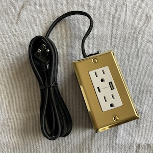 Peut inclure: Une prise électrique dorée avec deux prises standard et un port USB. Elle est dotée d'un cordon et d'une fiche noirs. Le couvercle de la prise est rectangulaire et fixé avec deux vis.