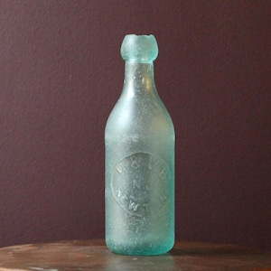 Puede incluir: Una botella vintage de vidrio transparente con un tono verde. La botella tiene un fondo redondeado y un cuello estrecho. La botella tiene una etiqueta que dice "Rock River" en un diseño circular.