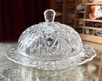 Op de afbeelding: Een glazen botervlootje van helder glas met een koepelvormig deksel en een geslepen glasontwerp. De schaal heeft een gegolfde rand en staat op een houten oppervlak.
