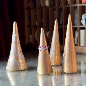 Puede incluir: Cuatro conos de anillo dorados con un anillo de piedra preciosa rosa y blanca en uno de los conos.