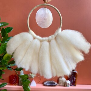 May include: White wool yarn wall hanging with a white seashell hanging from a wooden ring.
