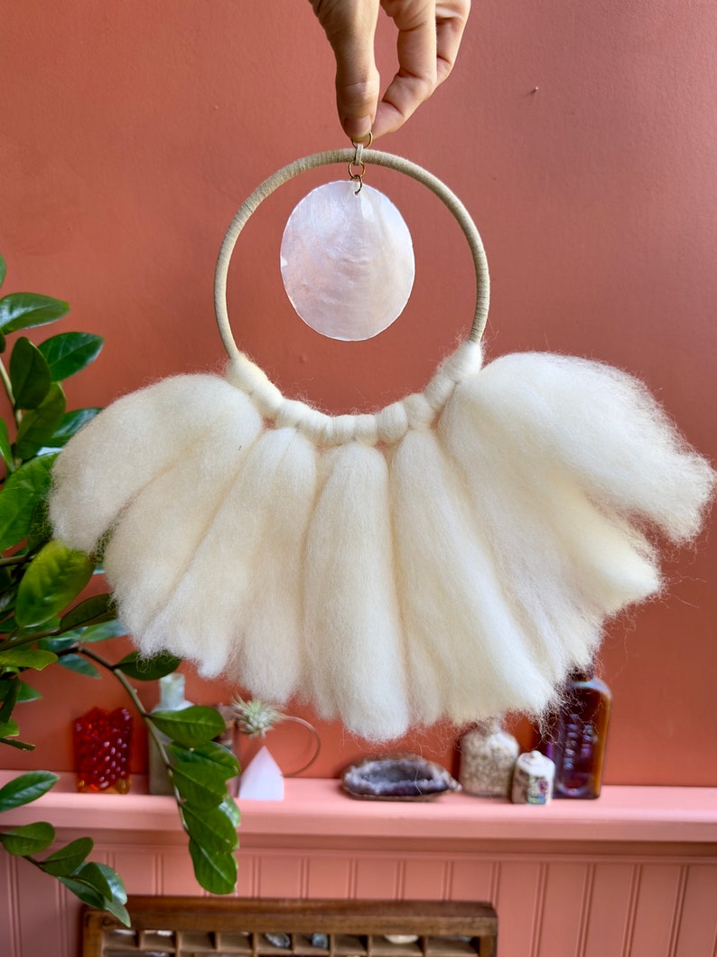 May include: White wool yarn wall hanging with a white seashell hanging from a wooden ring.