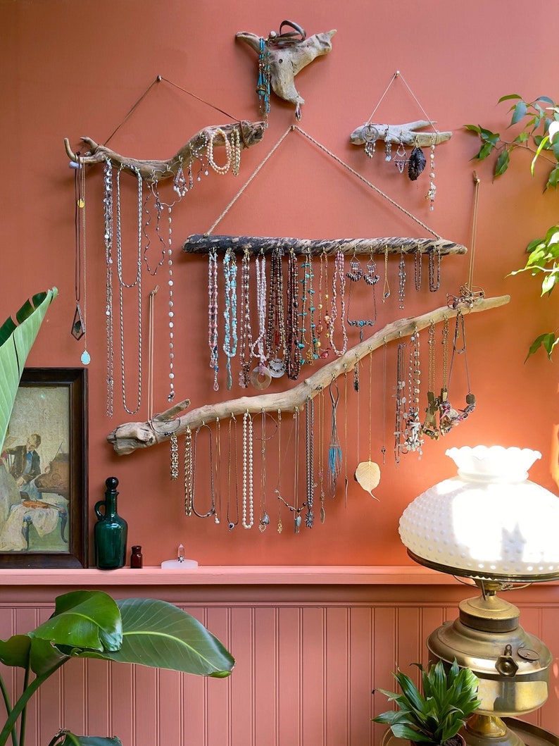 Puede incluir: Un organizador de joyas de pared hecho de ramas de madera flotante. El organizador presenta m&uacute;ltiples ramas colgantes y una rama horizontal larga. Las ramas est&aacute;n decoradas con collares, aretes y pulseras. La pared est&aacute; pintada de un color melocot&oacute;n claro.