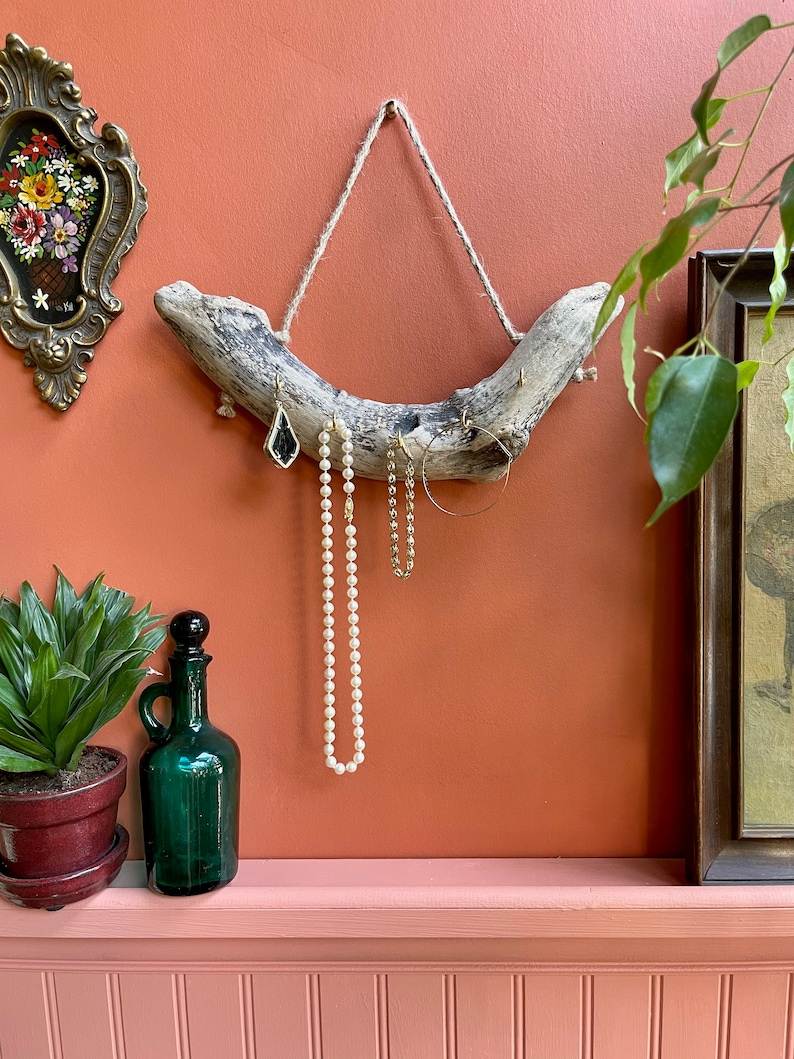 Puede incluir: Un soporte de joyer&iacute;a de madera flotante con un colgador de cuerda. El soporte tiene varios ganchos para colgar aretes, collares y pulseras. El soporte est&aacute; montado en una pared de color melocot&oacute;n.