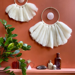 May include: Two white wool macrame wall hangings with a pink shell in the center of each circle. The hangings are on a terracotta wall with a shelf below. The shelf has a variety of decorative items, including a plant, bottles, and crystals.