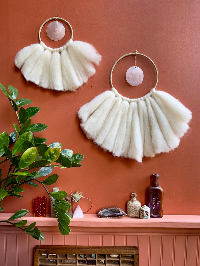 May include: Two white wool macrame wall hangings with a pink shell in the center of each circle. The hangings are on a terracotta wall with a shelf below. The shelf has a variety of decorative items, including a plant, bottles, and crystals.