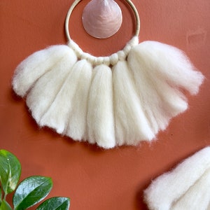 May include: White wool wall hanging with a circular wooden frame and a pink seashell in the center.