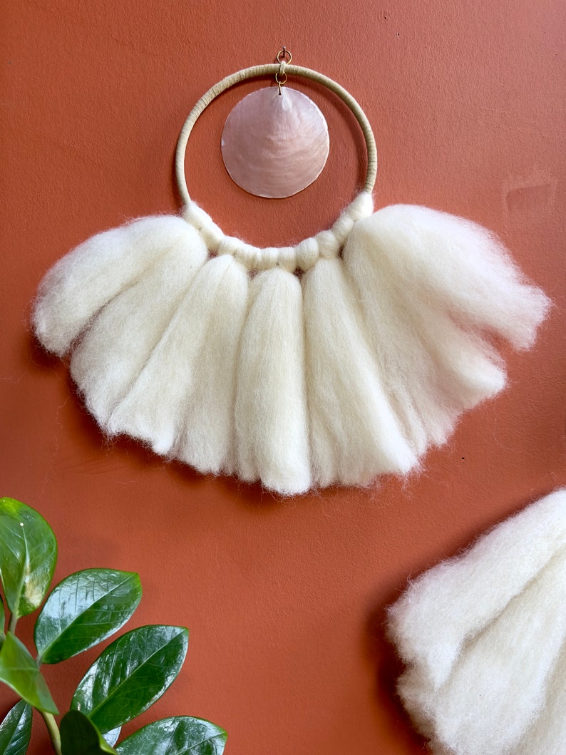 May include: White wool wall hanging with a circular wooden frame and a pink seashell in the center.