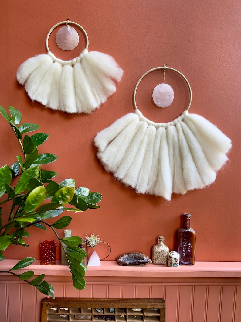 May include: Two white wool macrame wall hangings with a pink shell in each hoop. The hangings are on a terracotta wall with a shelf below. The shelf has a variety of decorative items, including a small plant, a bottle, and a few crystals.