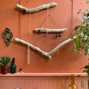 Puede incluir: Un organizador de joyas de madera flotante con tres ramas colgadas de la pared. Las ramas est&aacute;n decoradas con collares, pendientes y otras joyas. La pared est&aacute; pintada de un color naranja c&aacute;lido.